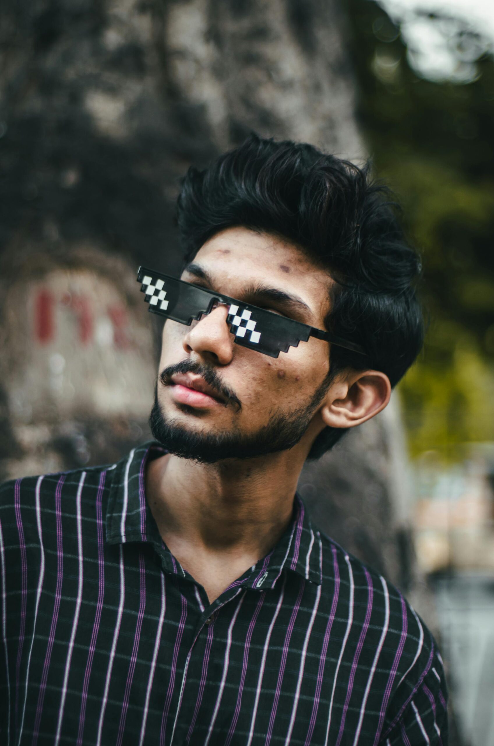 Hip bearded adult in pixel sunglasses and checkered shirt posing outdoors with a blurred background.