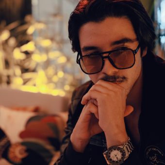 Stylish man with sunglasses sitting indoors, relaxed and contemplative.